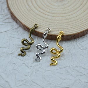 Puede incluir: Tres colgantes de metal con forma de serpiente en bronce, plata y oro. Cada colgante es una representación estilizada de una serpiente, con un lazo en la parte superior para sujetarlo a joyas u otras manualidades. Las serpientes están enroscadas y detalladas.