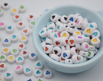 May include: A bowl filled with white round beads with colorful hearts printed on them. The beads are scattered around the bowl on a white surface.