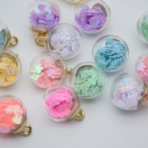 Op de afbeelding: Assortiment van kleine glazen ornamenten gevuld met kleurrijk, iriserend confetti. Elk ornament heeft een gouden lus om op te hangen.