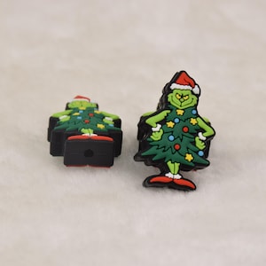 May include: Two Christmas-themed shoe charms. One charm is a green Grinch character wearing a Santa hat and a Christmas tree outfit. The other charm is a Grinch character in a Christmas tree outfit. The charms have red, green, yellow, and blue accents.