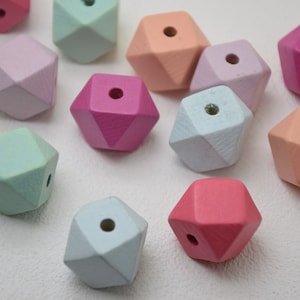 Może przedstawiać: Zbiór geometrycznych drewnianych koralików w pastelowych kolorach z otworem w środku każdego koralika. Koraliki są w różnych odcieniach różowego, niebieskiego i zielonego.