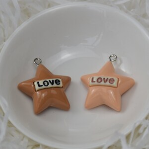 May include: Two star-shaped charms in shades of brown and peach, each with the word "Love" in a white rectangle. The charms have a silver loop at the top for hanging. They are displayed in a white bowl.