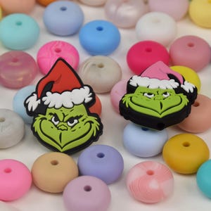 May include: A selection of colourful silicone beads in shades of pink, blue, yellow, and green. Two Grinch-themed silicone charms with Santa hats are also present. These beads are suitable for crafting and jewellery making.
