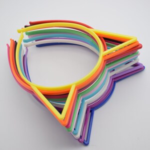 K&ouml;nnte beinhalten: Ein Stapel bunter Plastik-Haarreifen mit Katzenohren-Designs. Die Haarreifen sind in verschiedenen Farben, darunter Rot, Orange, Gelb, Gr&uuml;n, Blau, Lila und Wei&szlig;.