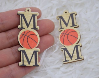 Könnte beinhalten: Holzanhänger mit dem Wort "MOM" in schwarzen Buchstaben, über und unter einem Basketball-Motiv. Der Anhänger ist hellbraun und hat oben ein kleines Loch zum Aufhängen.