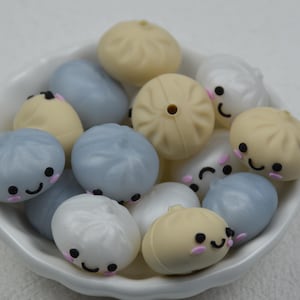 Peut inclure: Une collection de petites perles rondes en forme de raviolis, dans des tons beige, blanc et bleu clair, contenues dans un bol blanc. Chaque perle présente un visage souriant imprimé avec des yeux noirs, des joues roses et un trou central.