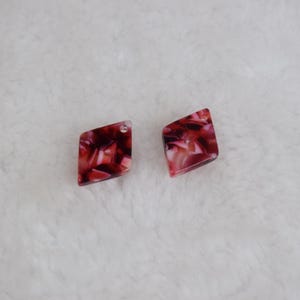 Puede incluir: Dos pendientes geométricos con un patrón veteado en tonos rojos y rosas. Cada pendiente tiene forma de rombo con un pequeño agujero para la fijación. Los pendientes están sobre un fondo blanco texturizado.