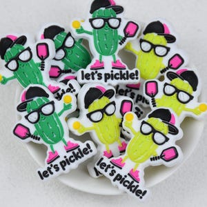 Peut inclure: Plusieurs breloques de cornichons de dessin animé, chacune portant des lunettes de soleil, une casquette de baseball et des baskets roses, tenant une raquette de pickleball. Le texte "let's pickle!" est imprimé sur chaque breloque.