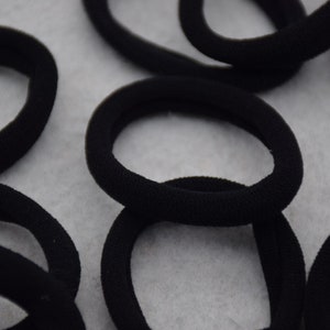 May include: A close-up of multiple black hair ties, made of a soft, stretchy material. The hair ties are arranged in a random pattern on a white surface.