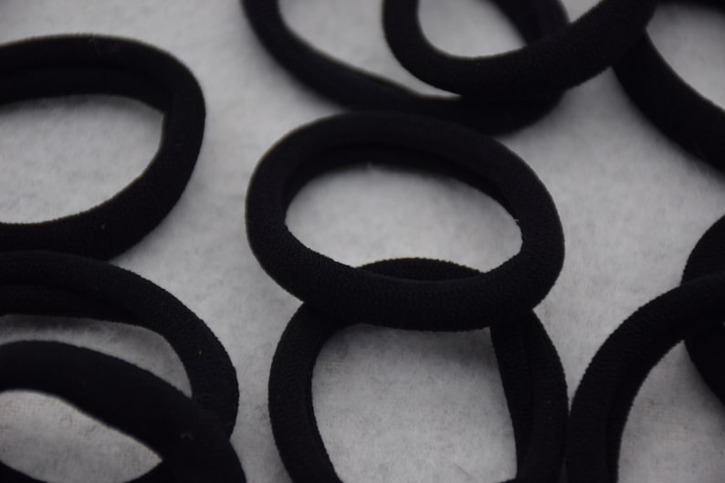 May include: A close-up of multiple black hair ties, made of a soft, stretchy material. The hair ties are arranged in a random pattern on a white surface.
