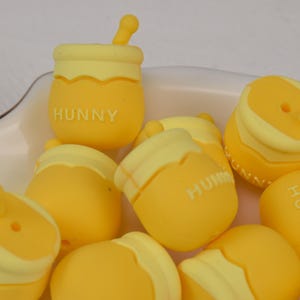 May include: A collection of yellow silicone honey jar beads with a light yellow rim and the word "Hunny" printed on each bead. The beads are designed to look like honey jars with a small yellow stick on top.