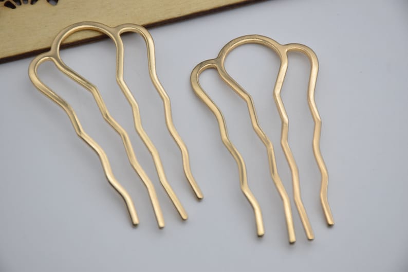 Op de afbeelding: Twee gouden haar kammen met een golvend ontwerp. De kammen hebben elk drie tanden.