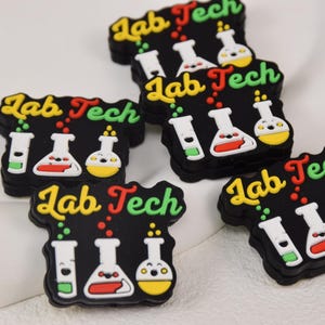 Puede incluir: Cinco dijes de silicona negros y coloridos con el texto "Lab Tech" y un gráfico de vasos de precipitados y tubos de ensayo.