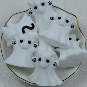 May include: A collection of white ghost-shaped figurines with black paw prints and eyes, arranged in a white bowl with a gold rim. One ghost has a black question mark. The figurines have a cartoonish design, perfect for Halloween decorations.