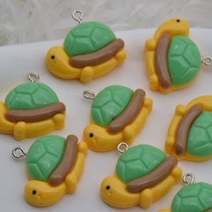 Puede incluir: Un conjunto de abalorios con forma de tortuga en amarillo, verde y marrón. Cada abalorio presenta un cuerpo amarillo, un caparazón verde y un elemento marrón en forma de tronco. Tienen un pequeño lazo plateado para sujetar.