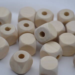 以下が含まれることがあります： 未完成の木製ビーズのコレクション。ビーズは立方体型で、角が丸く、中央に穴が開いています。天然木の色は薄いベージュです。