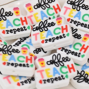 以下が含まれることがあります： 白のシリコン製チャームで、黒い文字とカラフルな文字で「coffee teach repeat」と書かれています。チャームには小さなコーヒーカップのアイコンが付いています。
