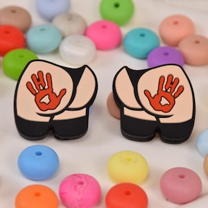 May include: Two novelty butt-shaped charms with a handprint design in red and tan, surrounded by colorful beads in various shades. The charms have black bottoms and are likely for crafting or jewelry making.