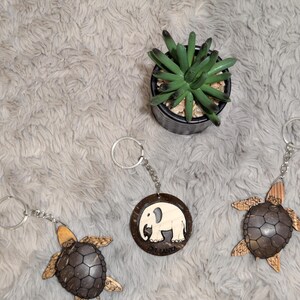 May include: Keyrings with turtle and elephant designs. The turtle keyrings have brown shells and tan flippers. The elephant keyring features a white elephant design on a brown background. A small potted succulent is also in the image.