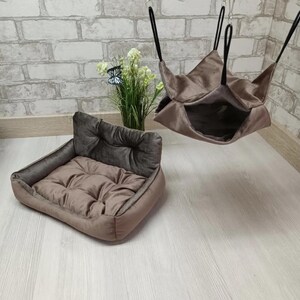 May include: Two pet beds: a rectangular, brown and grey plush bed with a tufted cushion and a hanging, star-shaped hammock. The hammock and bed are made of soft, velvety material. The background includes a brick wall and a vase of flowers.