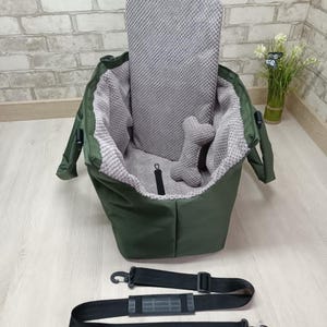May include: An olive green pet carrier bag featuring a textured grey interior lining, a matching grey upright back cushion, and a small bone-shaped grey toy. The carrier includes a detachable black adjustable shoulder strap with a padded section.