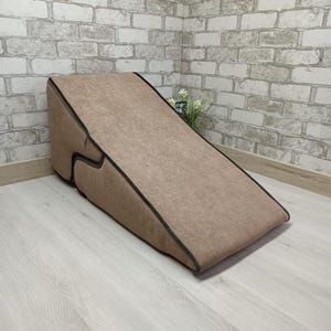 May include: A tan pet ramp with a dark brown trim. The ramp is angled and designed to help pets access elevated surfaces. The ramp is made of a soft, textured material and is placed on a light wood floor.