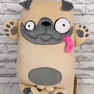 May include: A plush pug dog-shaped pillow with a beige body, gray accents, and a pink tongue. The pillow has large, wide eyes and paw-shaped details on the paws. It is sitting on a wooden box.
