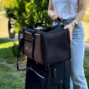 May include: A black rolling suitcase with a black and brown pet carrier on top. The pet carrier has a mesh front and a brown leather-like exterior. A black shoulder strap is attached to the pet carrier.