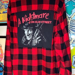 May include: Red and black plaid flannel shirt with a black patch featuring the text "A Nightmare on Elm Street" and a graphic of Freddy Krueger. The shirt is hanging on a wooden hanger.