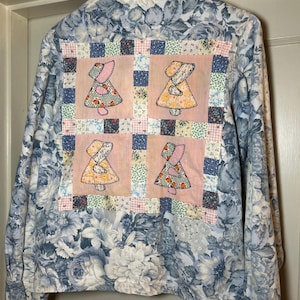 May include: A vintage button-up jacket with a floral pattern in shades of blue and white. The back features a patchwork design with four squares, each showcasing a figure in a dress. The jacket is hanging on a wooden hanger.