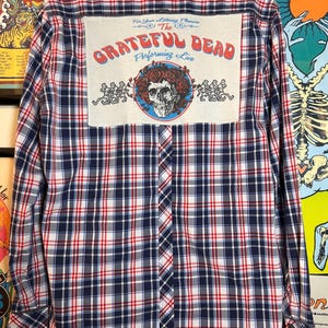 May include: A long-sleeved button-down shirt with a plaid pattern in red, white, and navy blue. The back features a patch with the text "Grateful Dead Performing Live" and a skull with roses graphic. A classic rock and roll shirt.