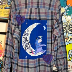 May include: Plaid button-down shirt in gray, blue, and burgundy. Features a blue patch with Eeyore from Winnie the Pooh on a crescent moon, with the text "I love you to the Moon and Back." Two purple heart patches are also visible.