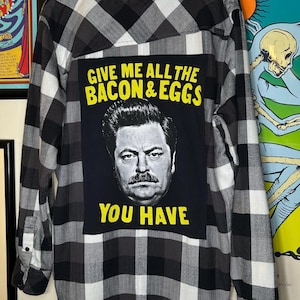 May include: A black, white, and grey checked flannel shirt. The back features a black square with a man's portrait and the yellow text: "GIVE ME ALL THE BACON & EGGS YOU HAVE". The shirt has long sleeves.