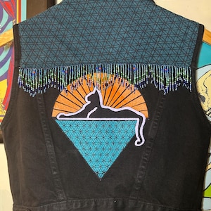 May include: Black denim vest with a decorative back panel featuring a cat silhouette against an orange sunburst. The vest has a turquoise geometric diamond, a teal patterned upper back, and beaded fringe.