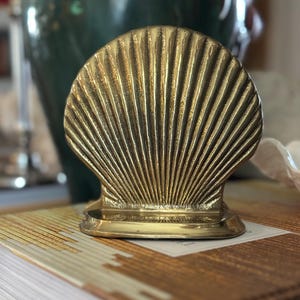 May include: A gold-colored, seashell-shaped bookend. The bookend has a fan-like design with vertical lines radiating from the base. It sits on a book with a patterned cover, with a blurred green vase in the background.