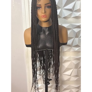 May include: A dark brown braided wig on a mannequin head. The wig features long, individual braids with a lace front. The mannequin is displayed on a black stand against a white textured wall.