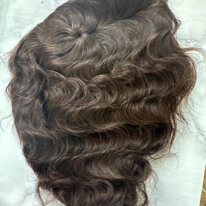 May include: A dark brown hair topper featuring a wavy texture and a visible crown section. The hair is styled with soft, flowing waves that cascade downward, showcasing a natural-looking part and a consistent deep brown color throughout the piece.