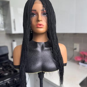 May include: A black braided wig on a mannequin head. The wig features small, tightly woven braids that cascade down the shoulders. The mannequin has a realistic face with makeup and is mounted on a black stand.