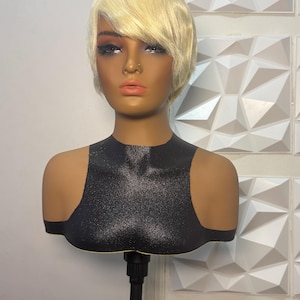 May include: A mannequin head with a short, blonde wig. The mannequin has a dark complexion, with makeup applied to the face. The mannequin is wearing a black, glittery top. The mannequin is mounted on a black stand.