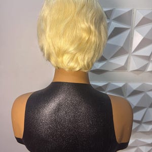 May include: A blonde wig with a short, wavy style is displayed on a mannequin head. The mannequin has a black top and a neutral skin tone. The background features a white geometric wall.