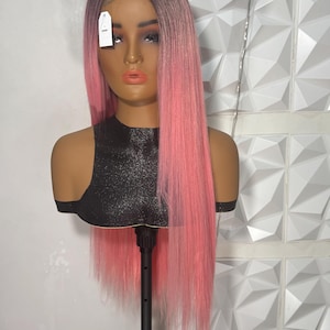 May include: A mannequin head displaying a long, straight wig. The wig transitions from a dark brown at the roots to a vibrant pink. The mannequin is on a black stand, and a tag hangs from the wig. The background is a white, textured wall.
