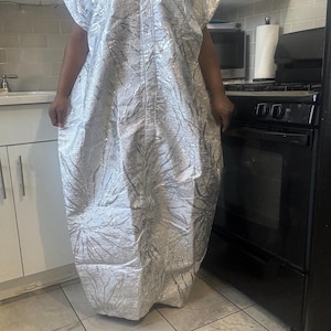 May include: A silver-colored, short-sleeved dress with a V-neck. The dress has a textured, metallic appearance with a subtle, leaf-like pattern. The dress is floor-length and appears to be made of a lightweight fabric.