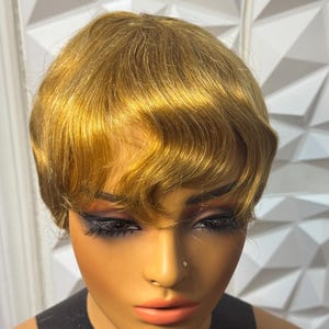 May include: A short, golden-blonde wig with a layered, wavy texture and soft bangs. The hair is styled in a cropped cut that frames the face, featuring natural-looking highlights and a smooth, polished finish throughout the entire head of hair.