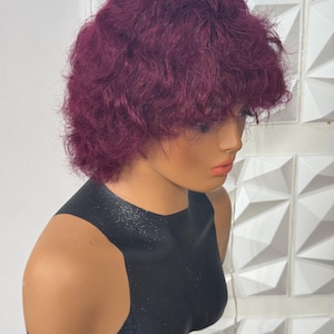 May include: A short, curly wig in a deep plum purple color with a textured, layered cut and bangs. The wig is displayed on a smooth, black mannequin head and shoulders, showcasing the vibrant hair color and the voluminous, wavy hair texture.