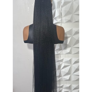May include: A long, black hair extension, likely a ponytail, is displayed on a mannequin head. The hair is straight and sleek, with a natural-looking texture. The mannequin is set against a white, textured wall.