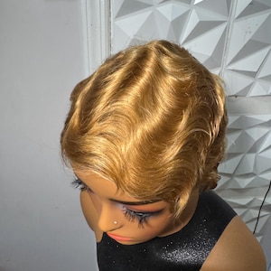 May include: A short, golden-blonde wig styled with deep, defined finger waves. The hair has a smooth, glossy texture and is fitted onto a mannequin head. The wig features a side part and follows a classic, short-cropped silhouette.