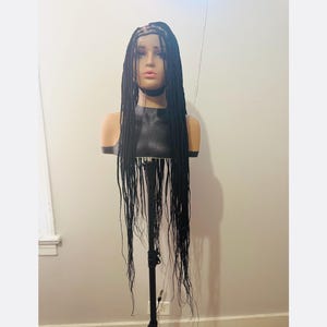 May include: A black braided wig displayed on a mannequin head. The wig features long, individual braids that cascade down. The mannequin is mounted on a black stand, set against a neutral wall.