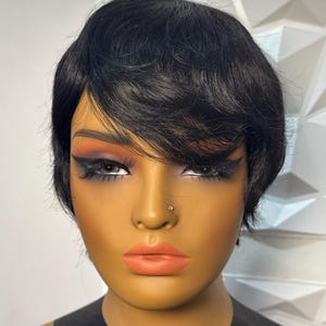 May include: A mannequin head with a short, black wig styled with side-swept bangs. The mannequin has a tan complexion, with expertly applied makeup including eyeshadow, eyeliner, and lipstick. The wig appears to be a pixie cut.
