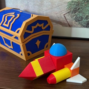 May include: A blue and gold treasure chest and a red, yellow, and blue toy rocket are displayed on a wooden surface. The rocket has a red nose cone and a blue dome on top.