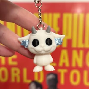 Può includere: Un portachiavi bianco con una piccola creatura dei cartoni animati con occhi neri, corna grigie e accenti blu sulle orecchie. Il portachiavi è attaccato a un anello e una catena di metallo argentato. Lo sfondo è un poster rosso sfocato.